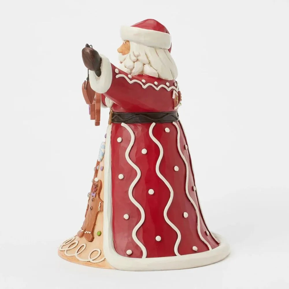 Side view of Santa figurine in red dotted robe with white wavy trim, holding wire cookie garland, gingerbread scene visible at hem
