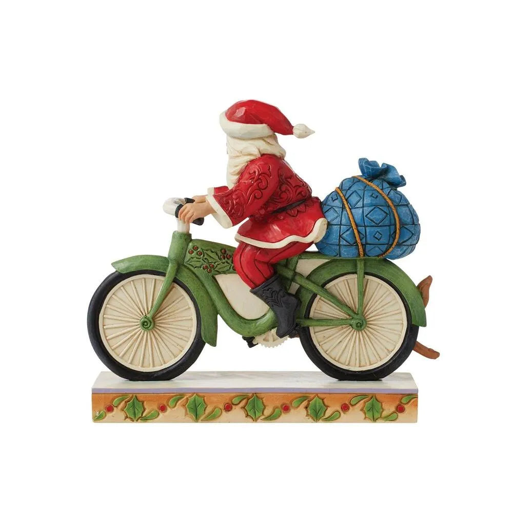 Rear view of Santa figurine cycling on green holly-detailed bicycle with blue gift sack secured to the back, holly base