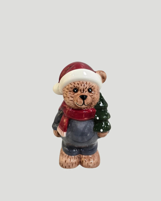 Ceramic teddy bear figure in santa hat and scarf holding a Christmas tree on plain background