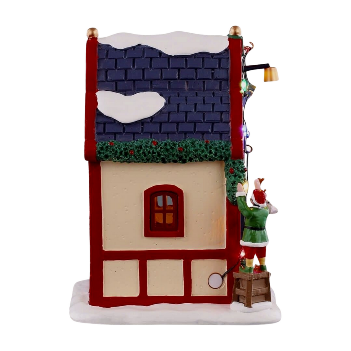 Right view of Comet's reindeer stable village building with blue roof, holly garland trim, lit window, and elf figurine on a crate