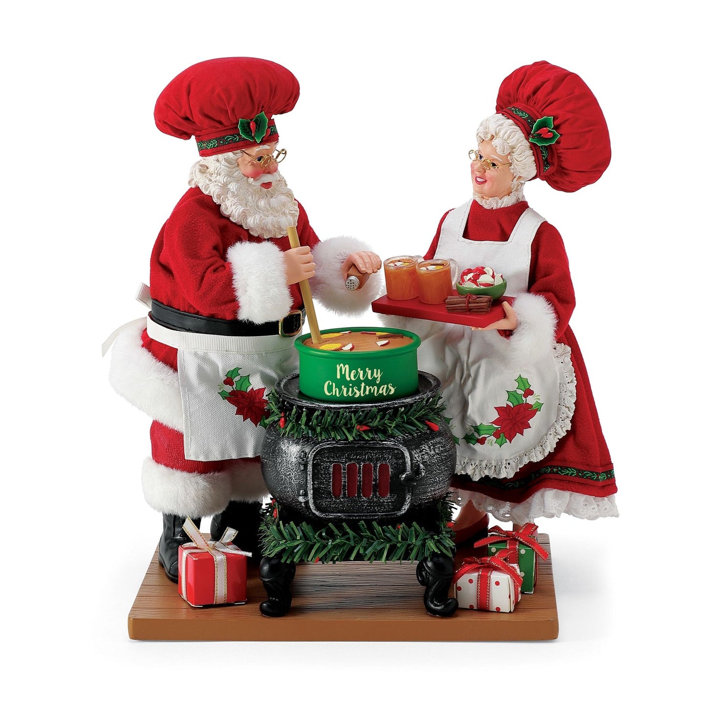 Santa and Mrs. Claus figurines in red chef outfits and holly-trimmed baker's hats, cooking at a garland-wrapped stove on a wooden base