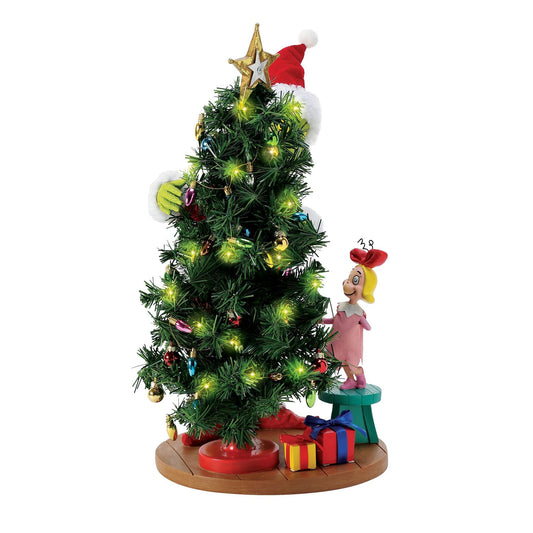 Side view of the Grinch figurine in a Santa suit at a lit Christmas tree, Cindy Lou Who figurine visible beside a teal stool 