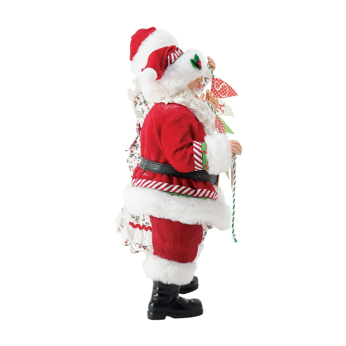 Side view of Santa figurine holding a "Merry Christmas" bunting banner, wearing a candy-stripe trimmed red suit with holly badge 