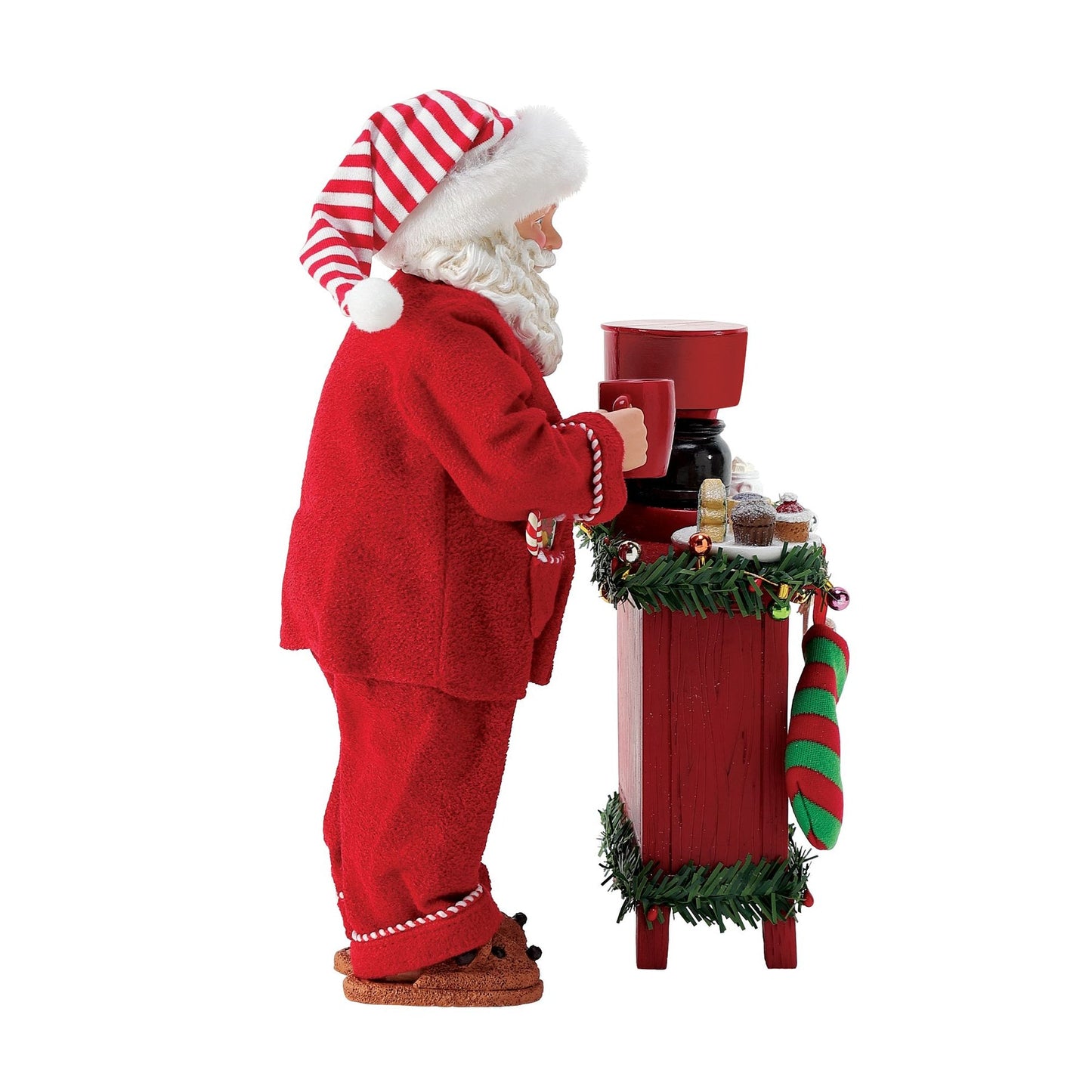 Side view of Santa figurine in red pyjamas and striped hat, holding a mug at a garland-decorated red sideboard with a coffee maker 