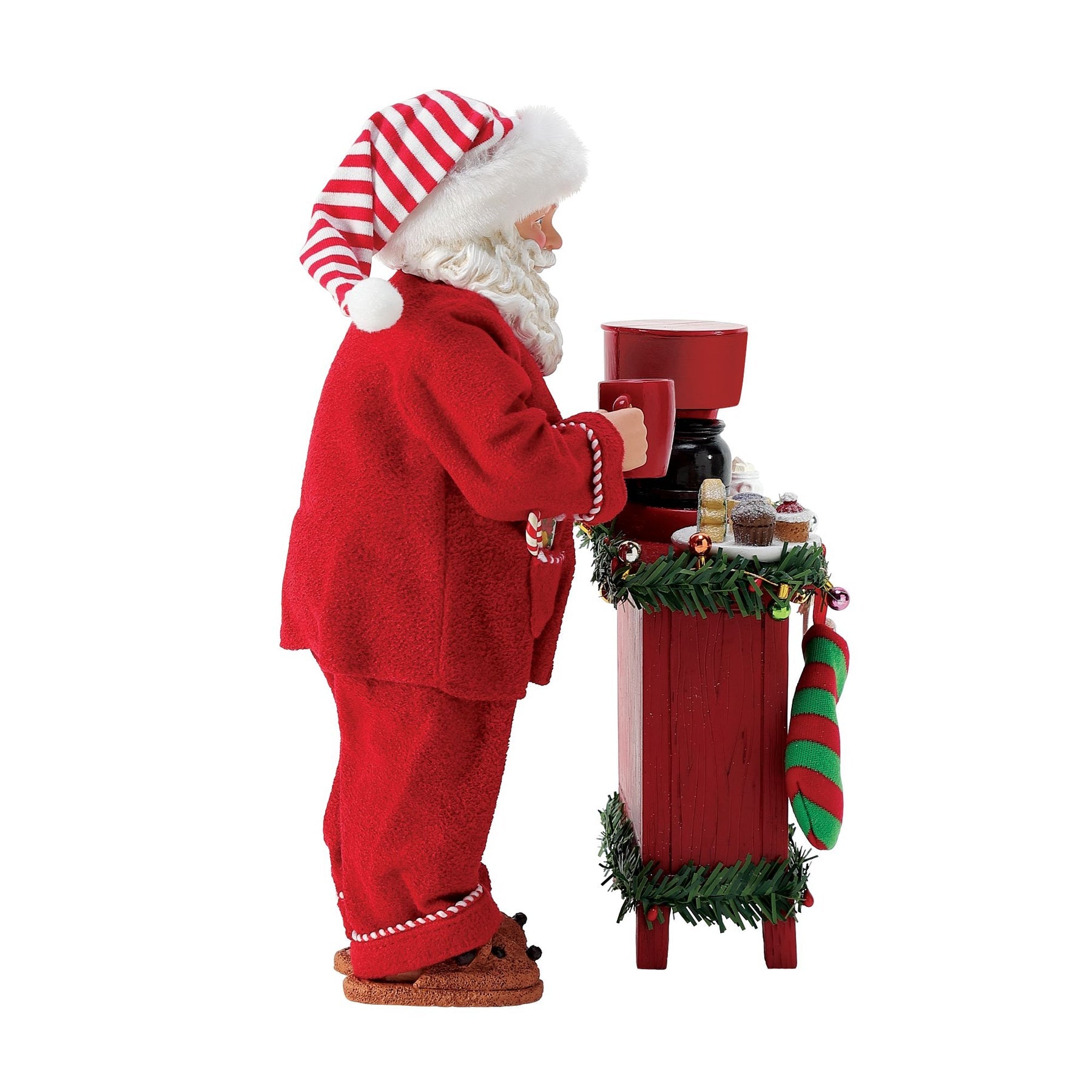Side view of Santa figurine in red pyjamas and striped hat, holding a mug at a garland-decorated red sideboard with a coffee maker 