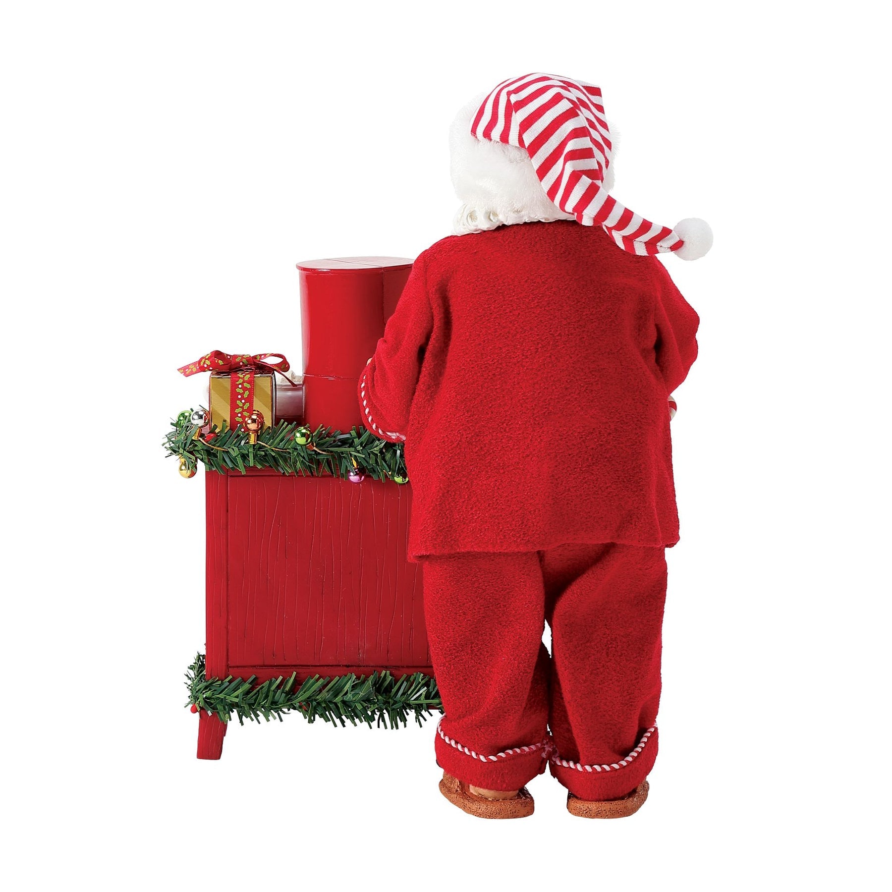 Rear view of Santa figurine in red pyjamas at a red sideboard decorated with garland, a wrapped gift, and a red coffee maker 
