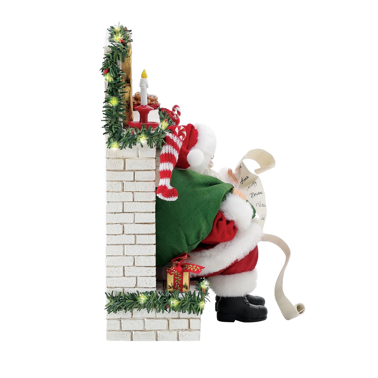 Left side view of Santa figurine seated on a lit garland-trimmed white brick fireplace, reading a long naughty-or-nice list, green sack beside him