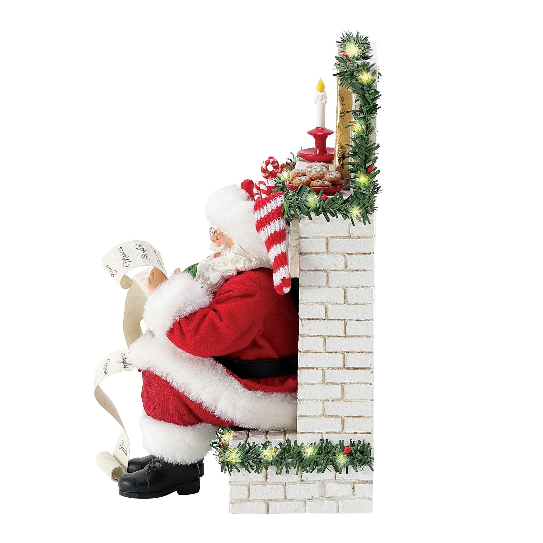 Right side view of Santa figurine seated on a garland-trimmed white brick fireplace reading his list, lit mantel with candle and cookies visible 