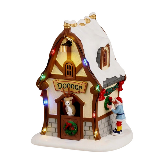 Side view of Donner's reindeer stable village building with snow-covered roof, red bow garland, wreath, and coloured lights