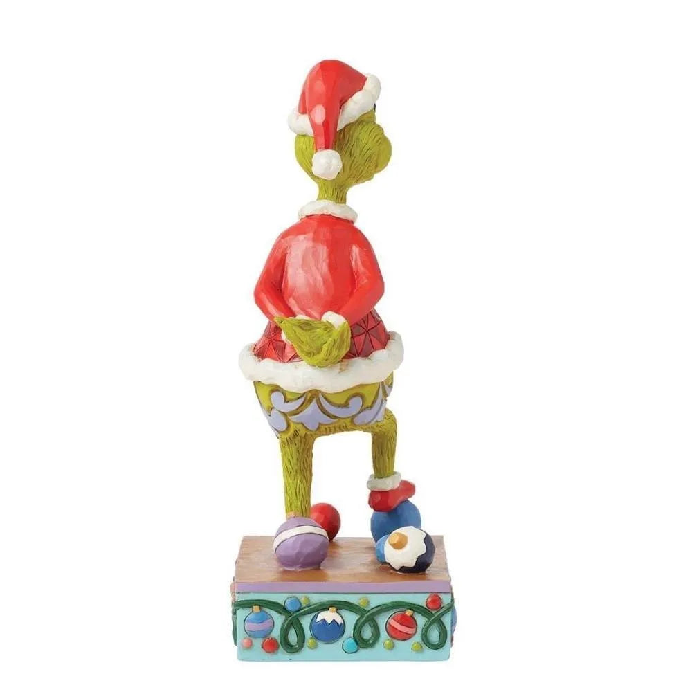 Side view of a Jim Shore Grinch Santa figurine with one leg raised, red geometric jacket, standing among scattered baubles on a decorative base