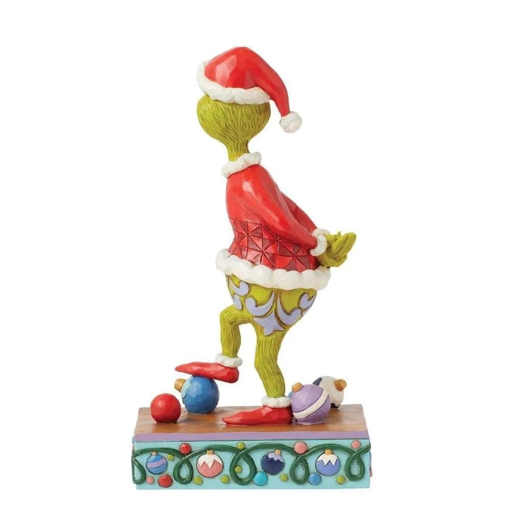 Rear view of a Jim Shore Grinch Santa figurine with hands clasped behind his back, scattered baubles at his feet, on a decorative base