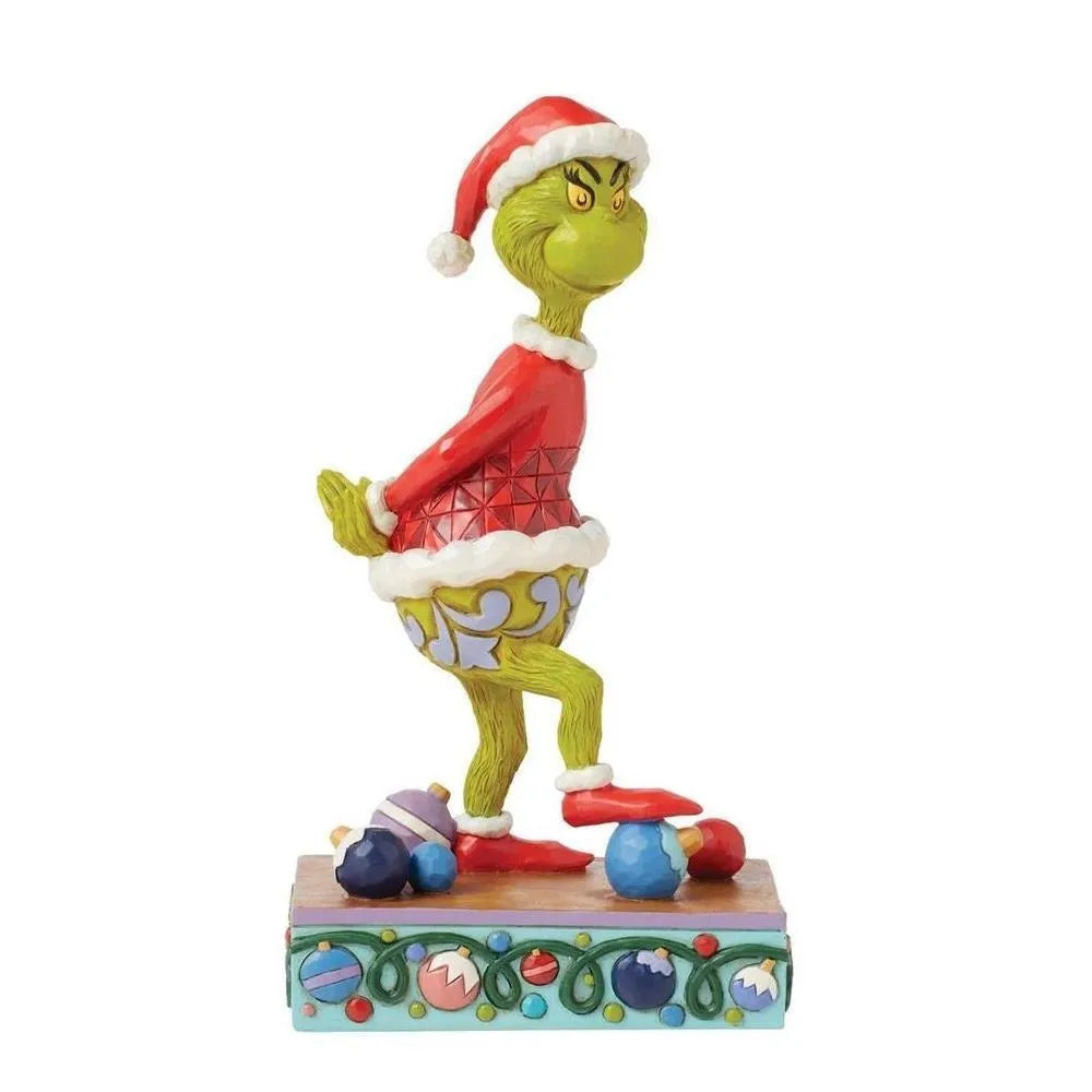 Angled front view of a Jim Shore Grinch Santa figurine with one leg raised, stepping on scattered baubles, on an ornament-detailed base. 