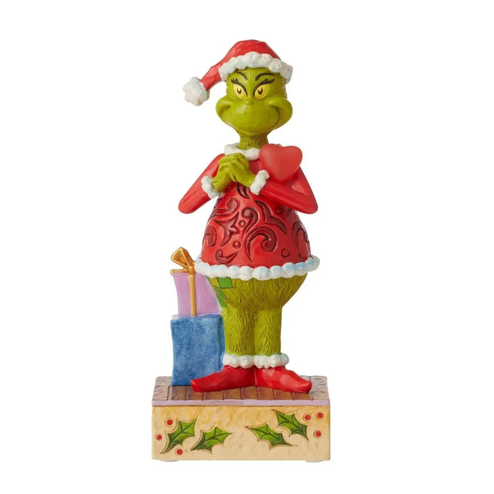 Grinch figurine in Santa hat and red scrollwork coat, clutching a heart, with stacked gifts beside a holly-decorated base