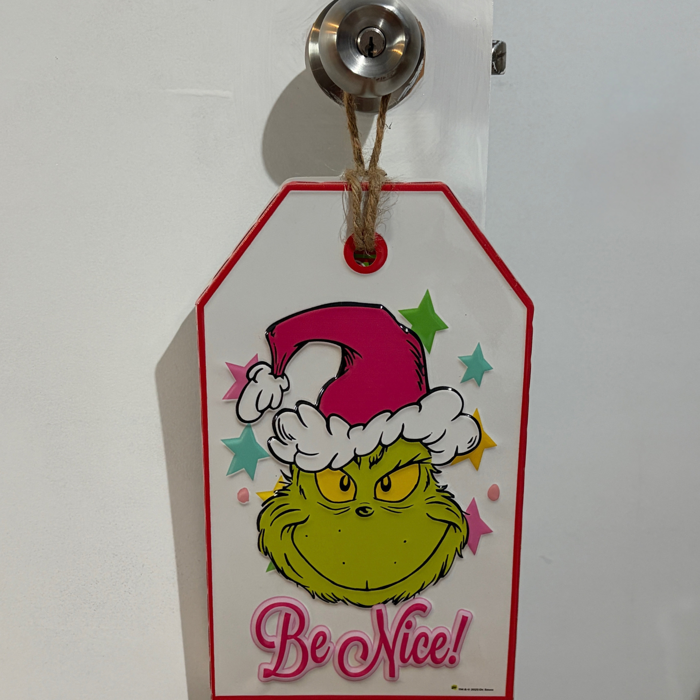 Door hanger featuring the Grinch wearing a Santa hat with colourful stars and the words “Be Nice!”