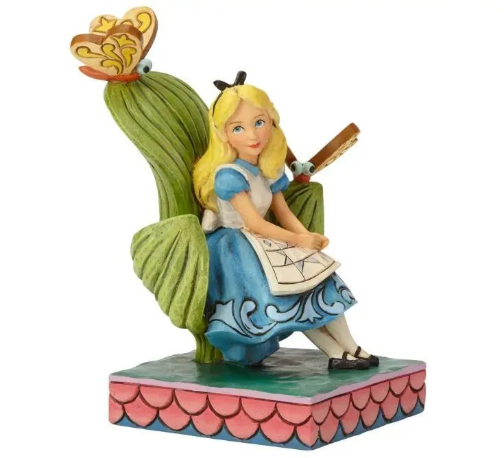Alice in Wonderland figurine side angle, seated with a book, flanked by large leaves and butterfly creatures on a pink scale base