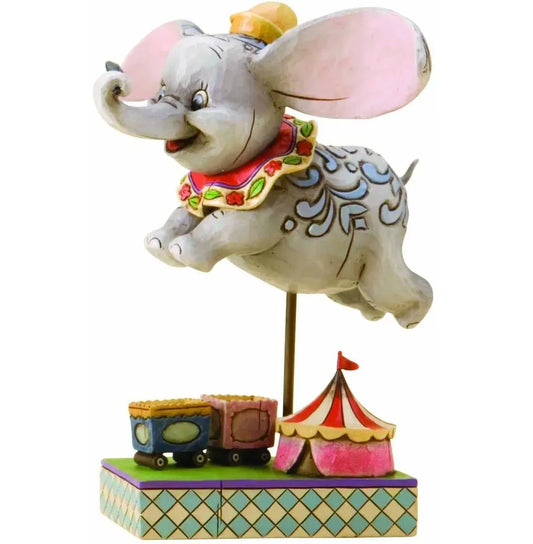 Dumbo figurine mid-flight above a painted circus tent and train cars base, wearing a floral collar with blue scrollwork detail 