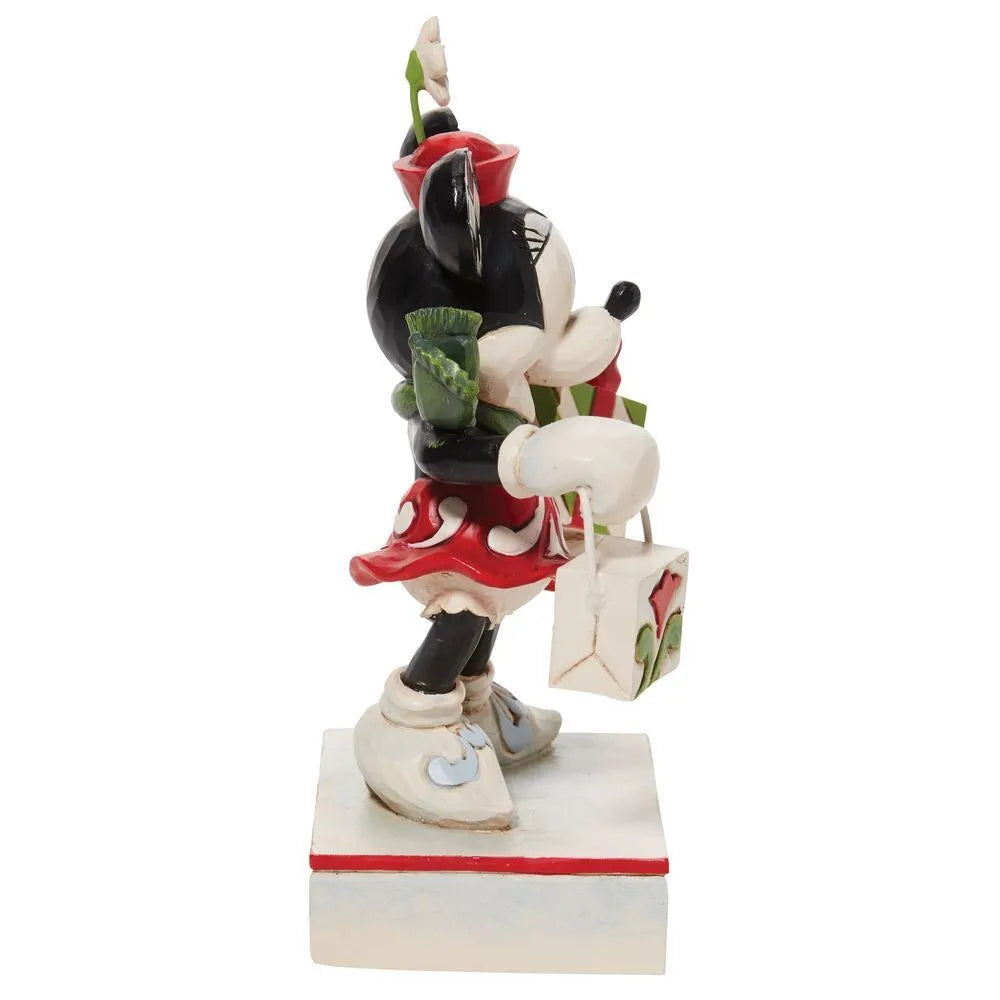Side view of Minnie Mouse figurine with checkered ears, red hat with flower, carrying a striped Christmas gift, holly base