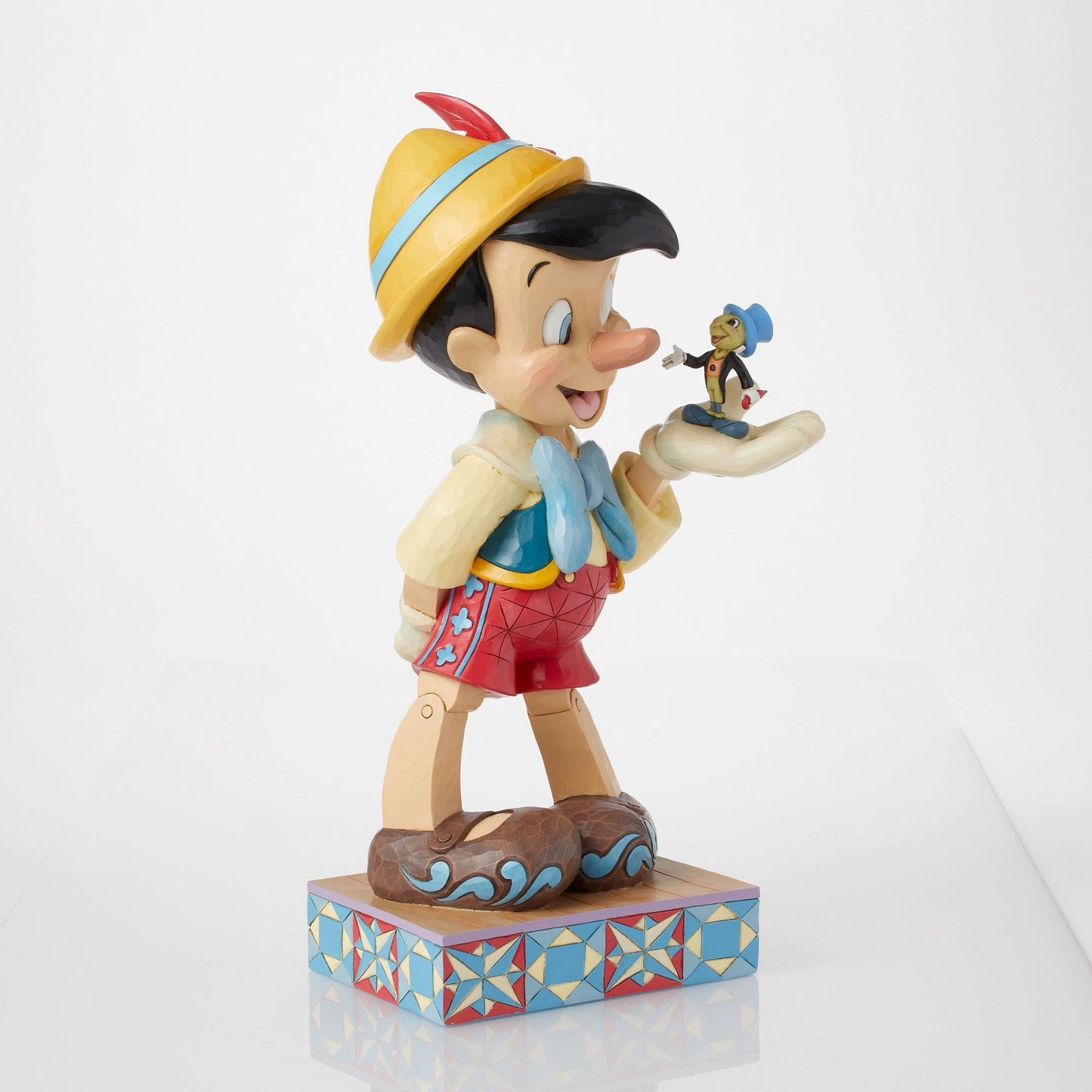 Front view of Pinocchio and Jiminy Cricket figurine, Pinocchio in yellow hat and red shorts, Jiminy on palm, star quilt base