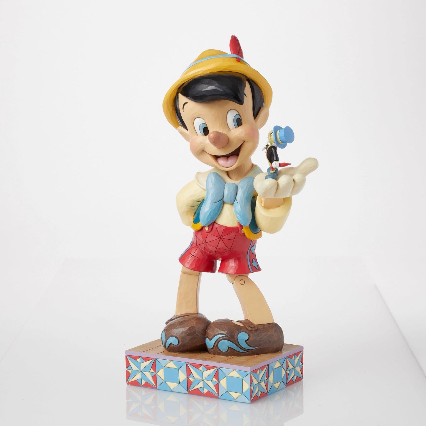 Right front angle of Pinocchio figurine holding Jiminy Cricket on palm, yellow hat with red feather, folk art star quilt base