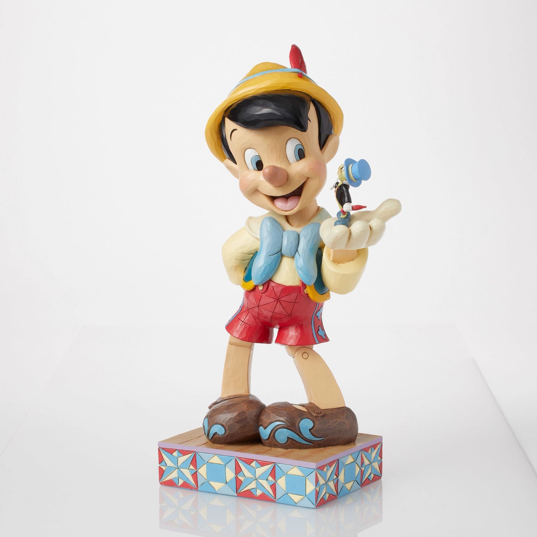Right front angle of Pinocchio figurine holding Jiminy Cricket on palm, yellow hat with red feather, folk art star quilt base