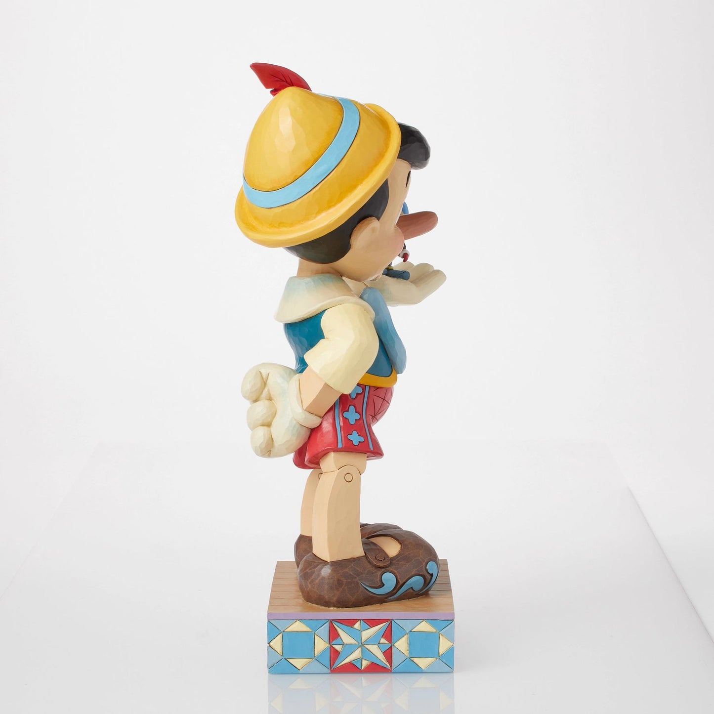 Left side view of Pinocchio figurine holding Jiminy Cricket on outstretched hand, yellow hat, red shorts, star quilt patterned base