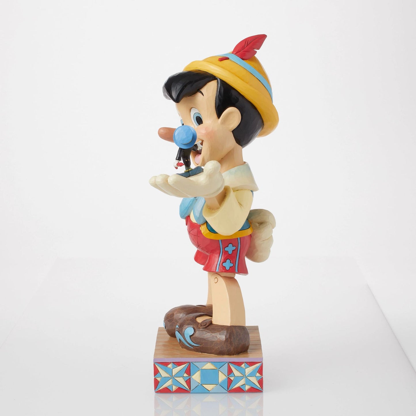 Right side view of Pinocchio figurine, yellow hat with red feather, blue waistcoat, Jiminy Cricket visible on outstretched hand
