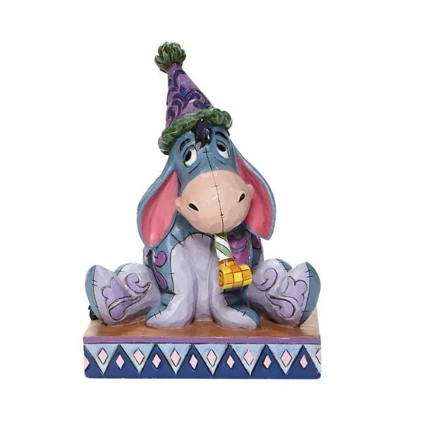Eeyore figurine in a party hat holding a blower, seated with a glum expression, on a diamond-patterned base