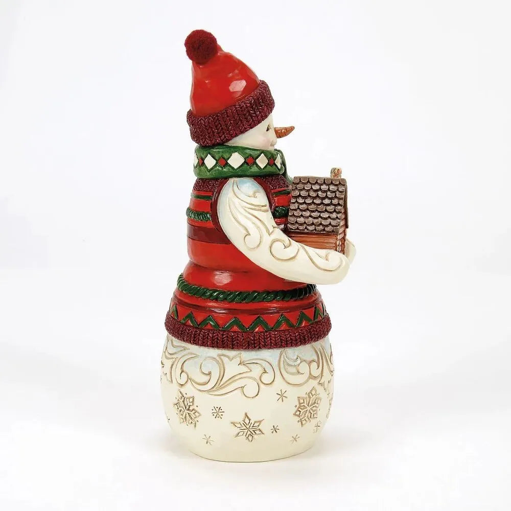 Side view of a Jim Shore snowman figurine holding a log cabin, wearing a red striped sweater and green argyle scarf, on a snowflake body