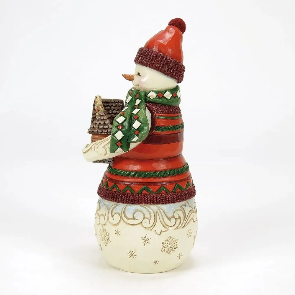 Opposite side view of a Jim Shore snowman figurine in a red bobble hat cradling a log cabin, with gold scrollwork snowflake lower body