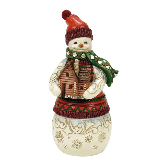 Front view of a Jim Shore snowman figurine in a red bobble hat and green argyle scarf, cradling a detailed log cabin
