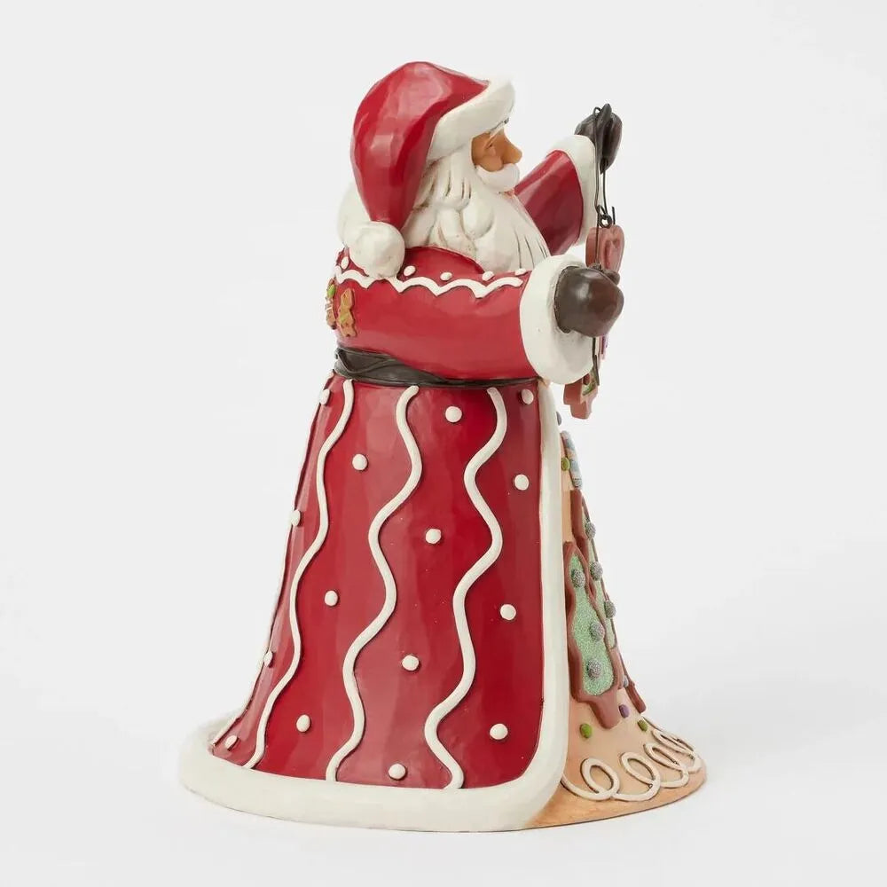 Left side view of Santa figurine in red robe with white wavy trim, holding dangling cookie ornaments, gingerbread scene on robe hem 