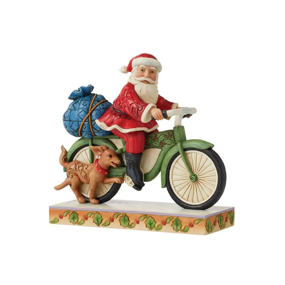 Side view of Santa figurine on green holly bicycle, blue gift sack on rear, small dog running beside, holly-decorated base