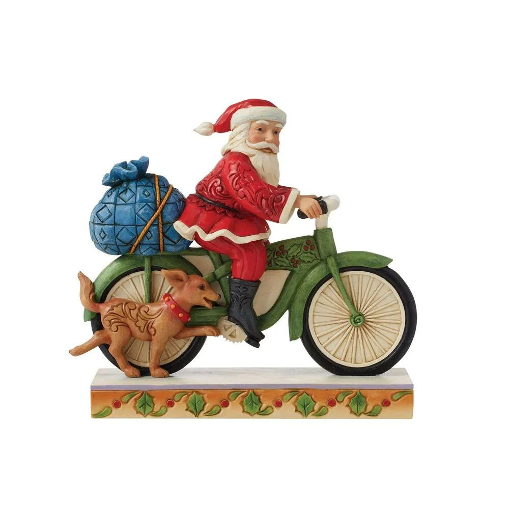 Santa figurine riding a green holly-detailed bicycle with a blue gift sack, a small dog running alongside, holly-decorated base