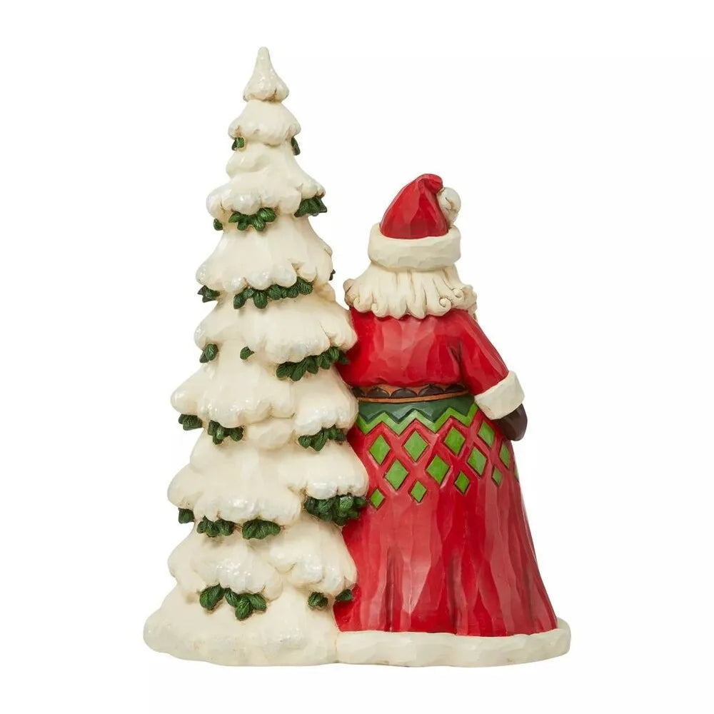 Rear view of Santa Claus figurine in red robe with green diamond pattern, beside a tall snow-covered folk art Christmas tree