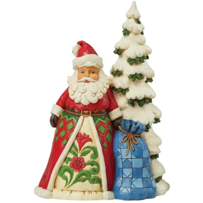 Santa Claus figurine in red folk art robe with floral scrollwork, standing beside a snow-covered tree and blue patchwork gift sack