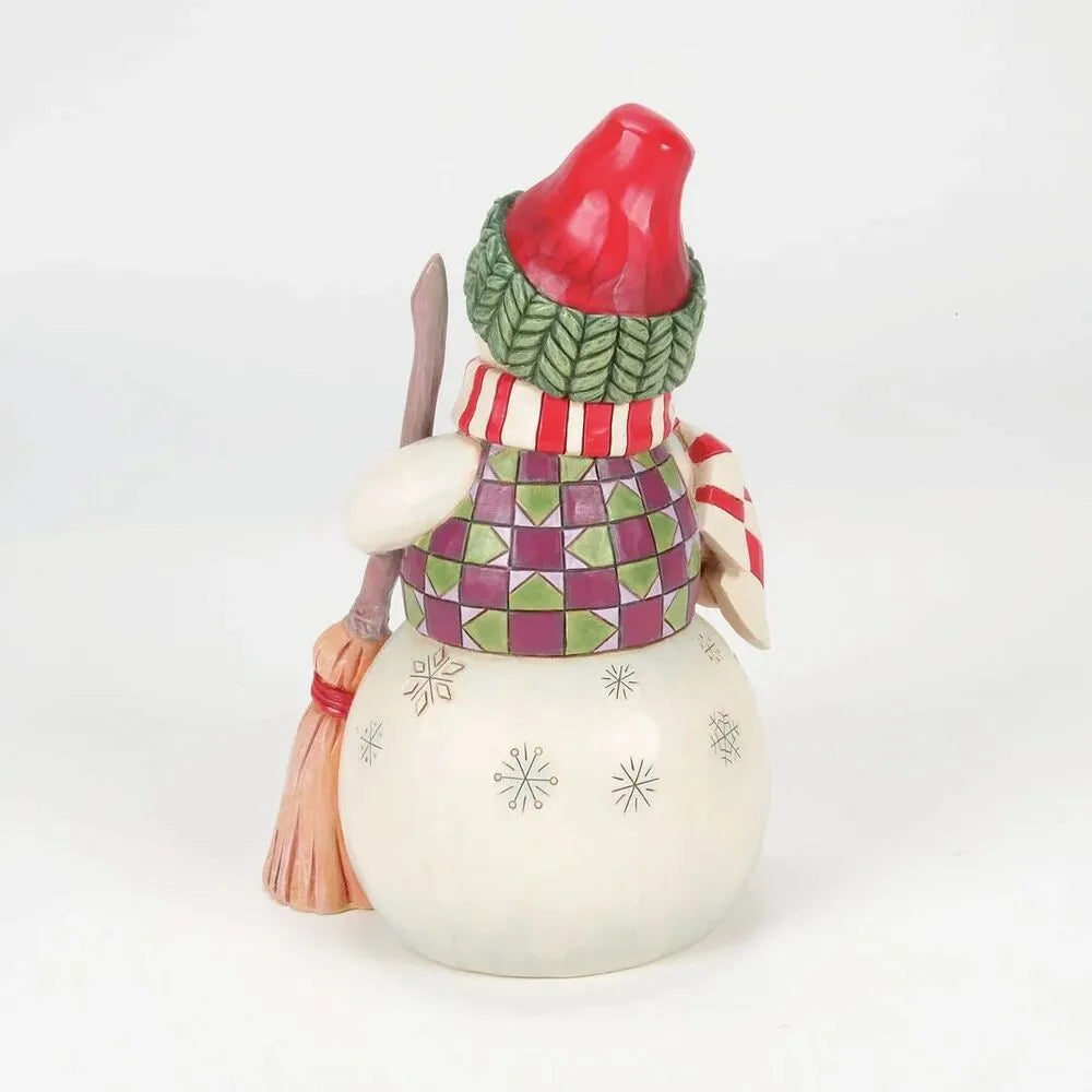 Rear view of a Jim Shore snowman figurine with red hat, green braided brim, diamond-pattern vest, striped scarf, and broom
