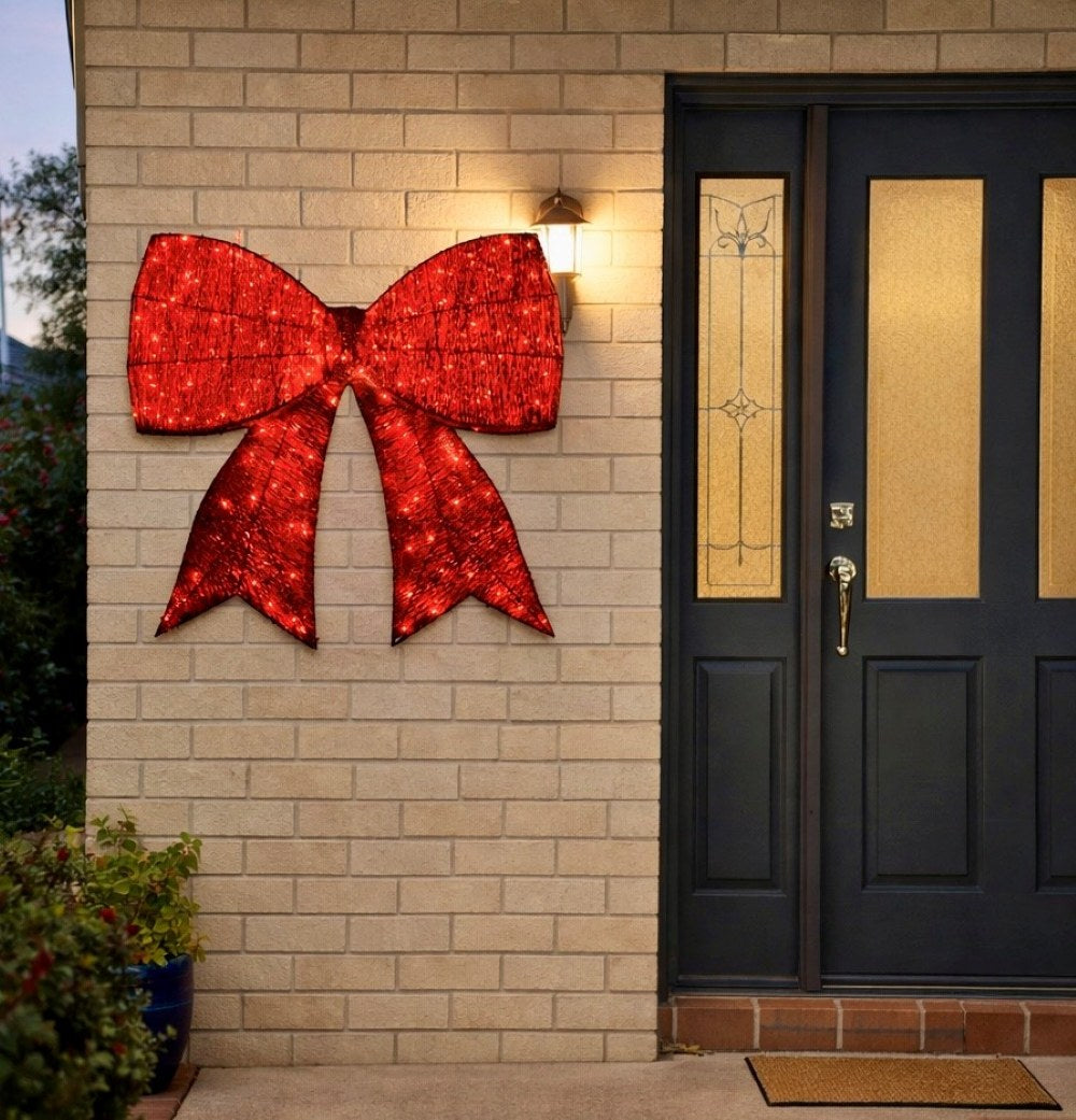 Large red LED light-up Christmas bow decoration mounted on exterior house wall beside front door
