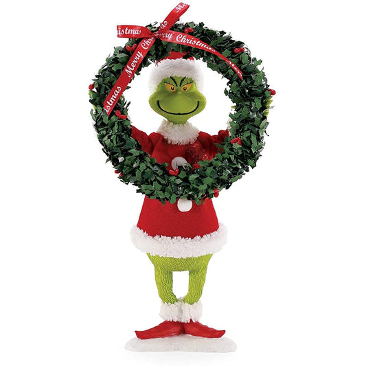 Grinch figurine in red Santa suit holding a large holly wreath with "Merry Christmas" ribbon, standing on a snowy base