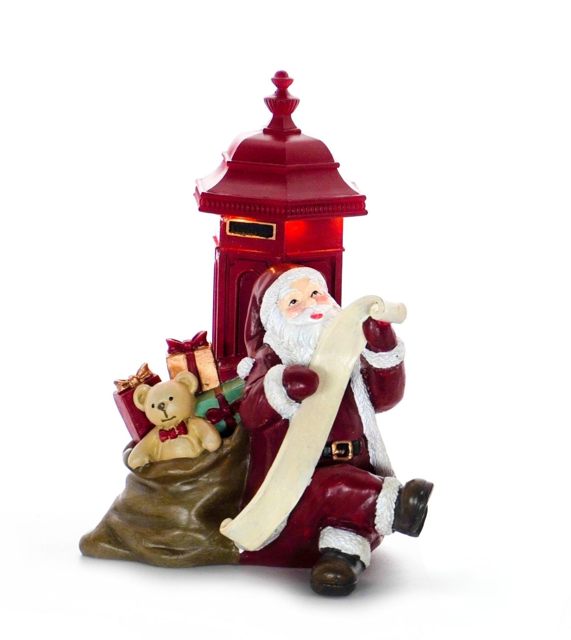 Santa figurine in red suit sitting beside a glowing red postbox with a sack of gifts and teddy bear while checking his list