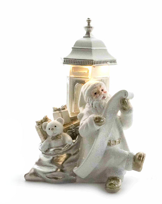 White and gold Santa figurine checking his list beside a glowing postbox with a sack of gifts and teddy bear