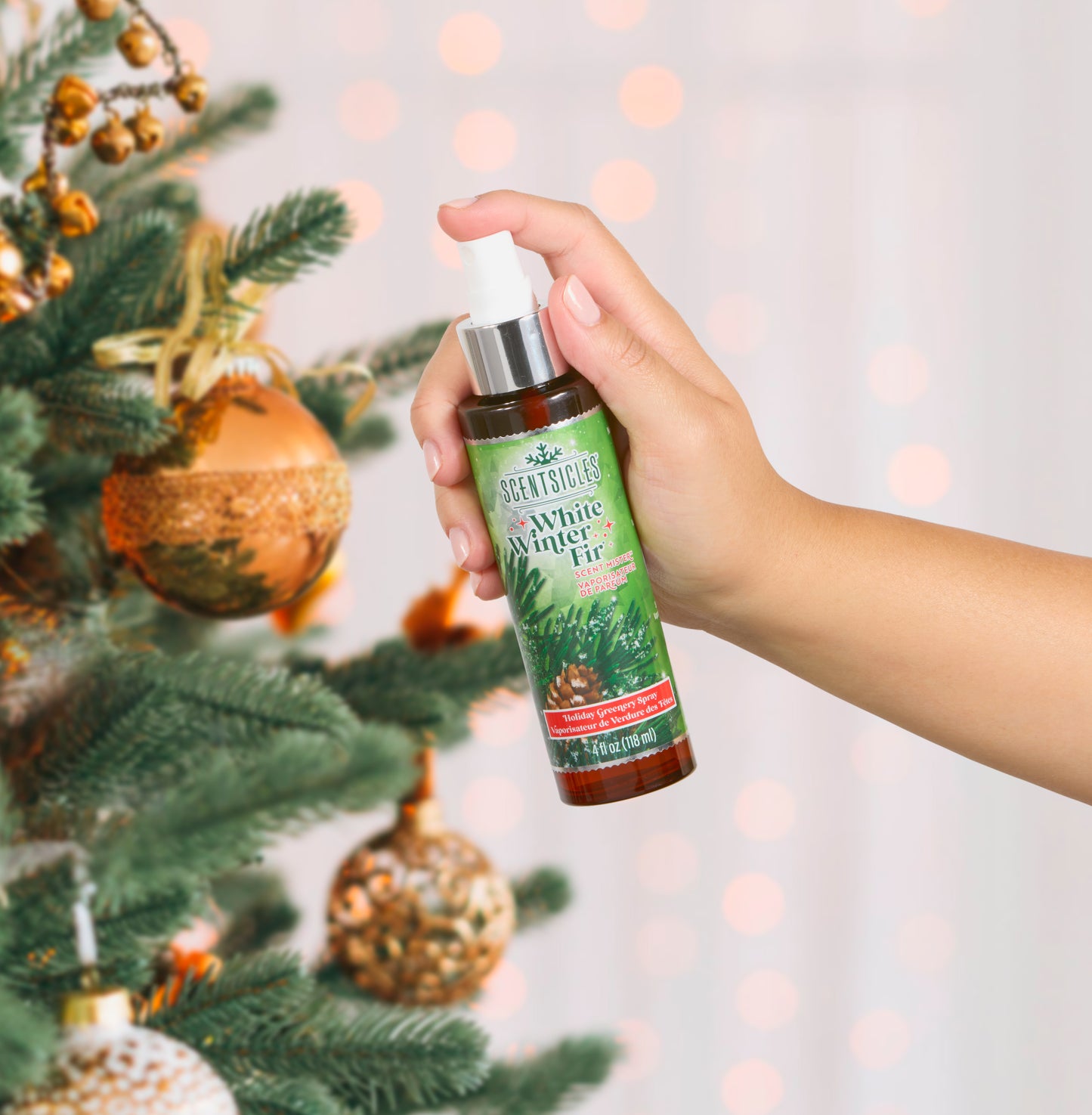 Hand holding a bottle of 'White Winter Fir' spray next to a decorated Christmas tree.