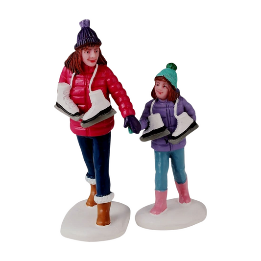 Set of two Christmas village figurines of a woman and girl in winter coats and beanies, each carrying ice skates, on snowy bases