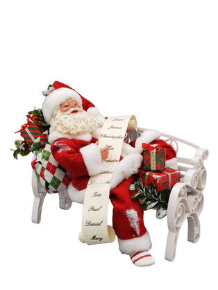 Santa Claus sleeping on a white bench holding a long list with gifts and greenery beside him