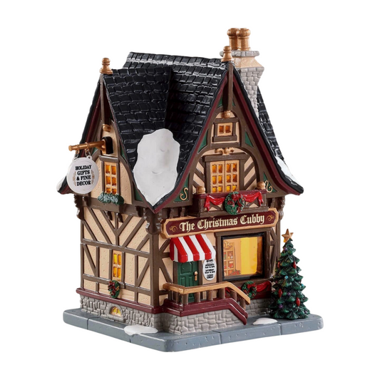 Lemax Christmas Cubby gift shop village building with Tudor-style exterior, striped awning, lit window display, and Christmas trees